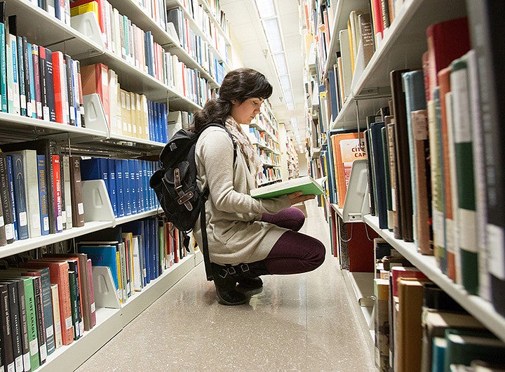 student research Student kneeling in a library aisle looking at an open book